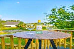 A round wooden table with two wine glasses and a bottle of white wine on a deck with a view of a grassy area and trees. - Luxe Dream Beach House (Gulfport)