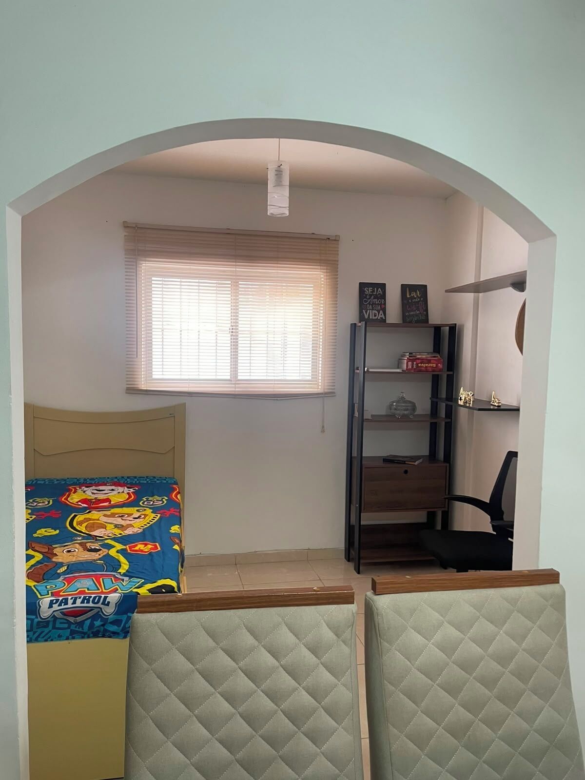 A room with a bed, a desk, a chair, a window with blinds, and a shelf with items.
