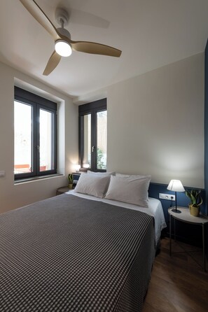 Ein Schlafzimmer mit einem Bett, zwei Fenstern, einem Deckenventilator, einem Nachttisch mit Lampe und einer kleinen Topfpflanze.