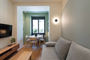 Ein Wohnzimmer mit einer Couch, einem Wandfernseher, einem Esstisch mit Stühlen und einer Topfpflanze.