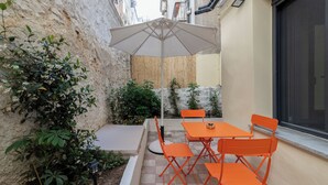 En udendørs terrasse med et bord, stole og en stor parasol, samt udsigt til en bygning.