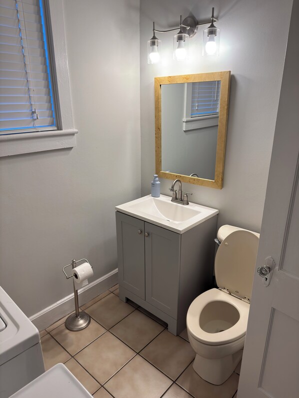Une salle de bain comprenant des toilettes, un lavabo, un meuble, un miroir et une fenêtre avec des stores.