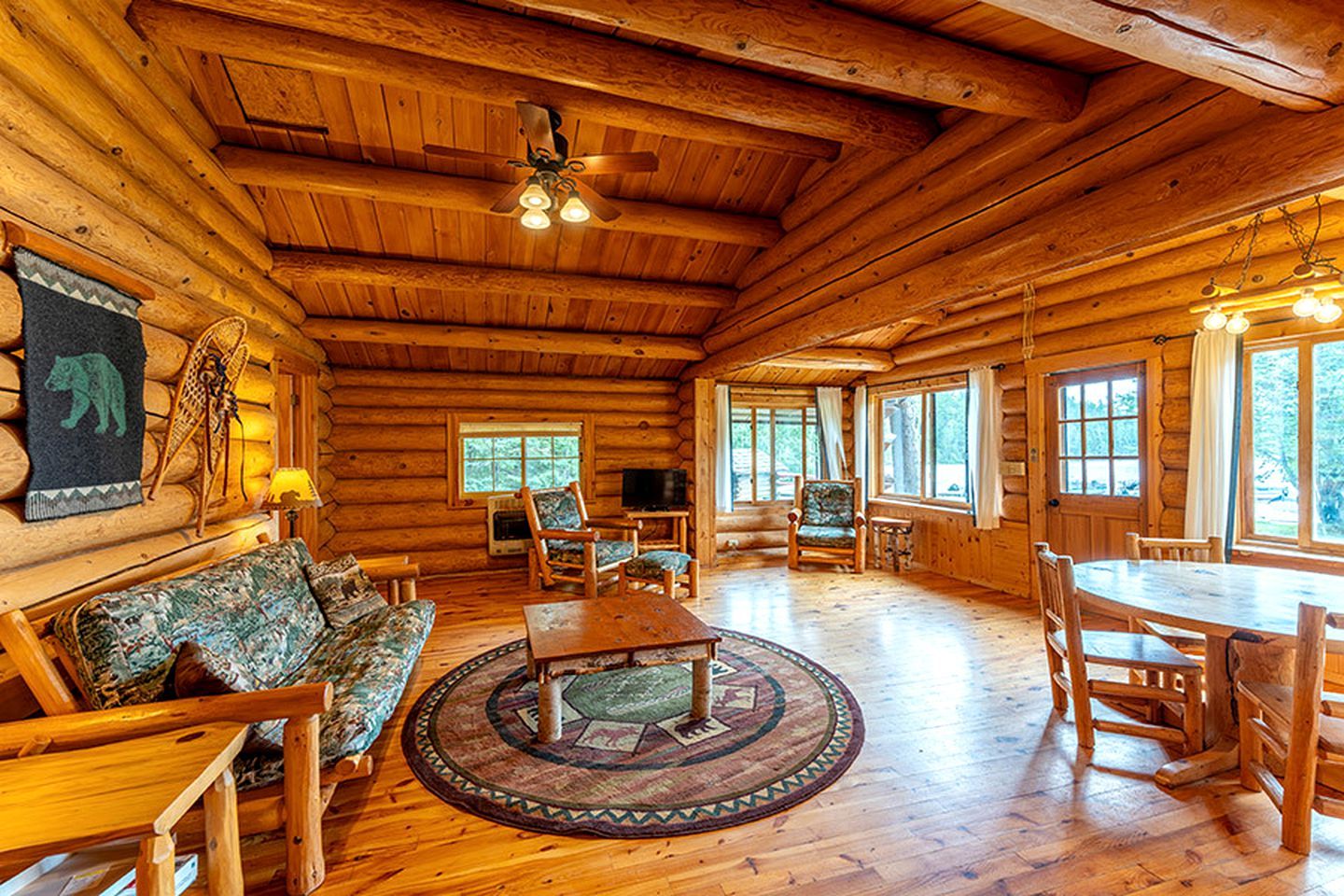 A log cabin interior with a wooden ceiling, a round coffee table, a sofa, a dining table with chairs, and a television.