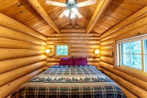 Ein Schlafzimmer in einer Blockhütte mit einem Bett, zwei Fenstern, einem Deckenventilator und Wandlampen.