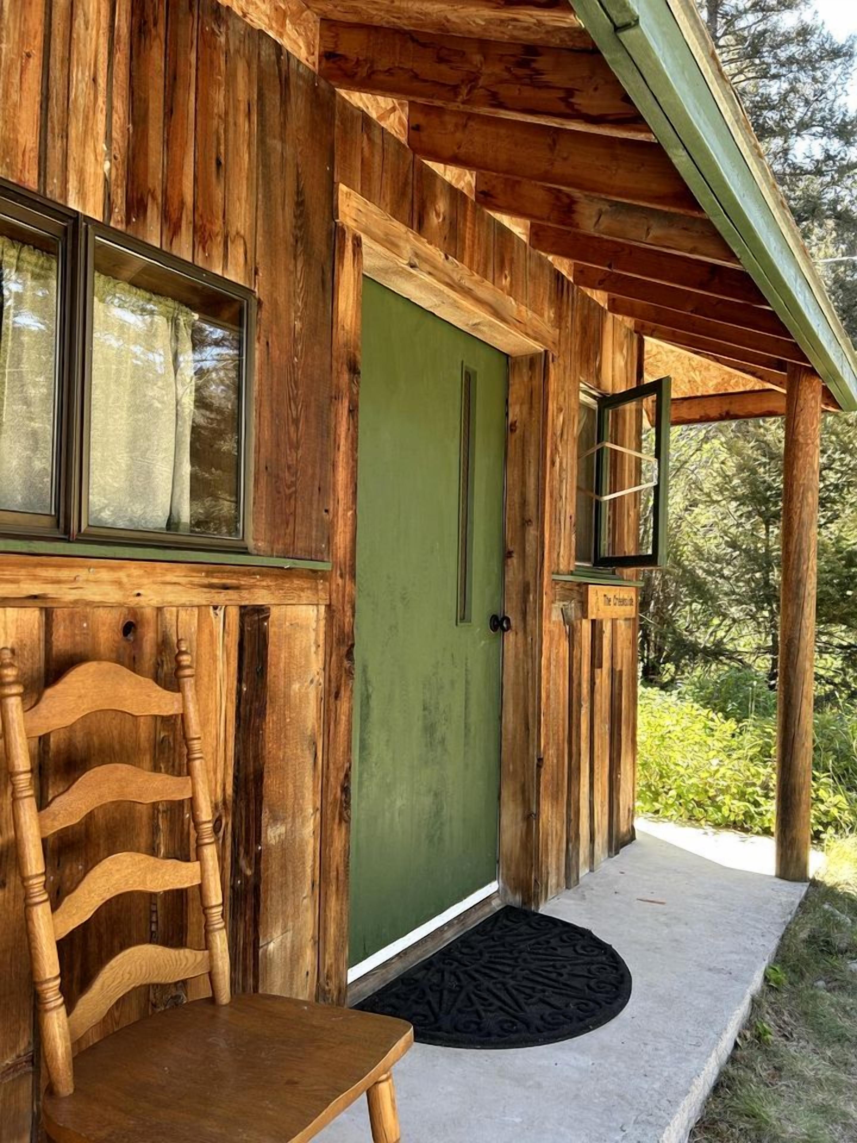 A wooden cabin with a green door, a window with curtains, a wooden chair, and a black doormat.