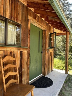 A wooden cabin with a green door, a window with curtains, a wooden chair, and a black doormat. - Secluded Family-Friendly Log Cabin Rental for Families near Helena (Hall)