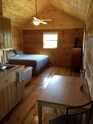 Quarto compacto no estilo de cabine, com cama, pequeno espaço de cozinha, mesa de jantar e ventilador de teto.