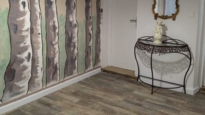 Une pièce avec un plancher en bois, une fresque murale représentant des arbres, une table ronde surmontée d’une statue et un luminaire suspendu.