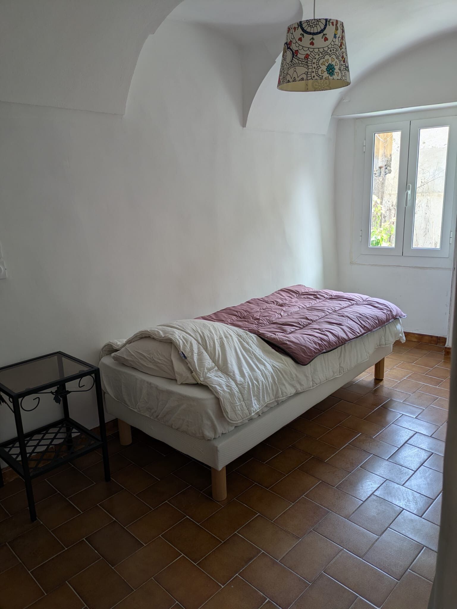 A single bed with a white and purple bedspread in a room with white walls, a window, and a tiled floor.