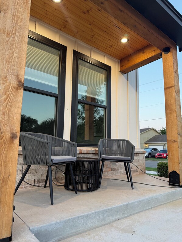 A covered patio with two chairs and a table, wooden beams, and a view of a street with parked cars.