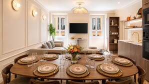 Een eetkamer met een grote houten tafel voor acht personen, een bank, een televisie en een keuken met houten kastjes en een betegelde achterwand.
