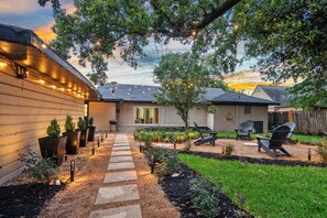 Un patio trasero con un sendero de piedra, mobiliario exterior e iluminación con luces colgantes.