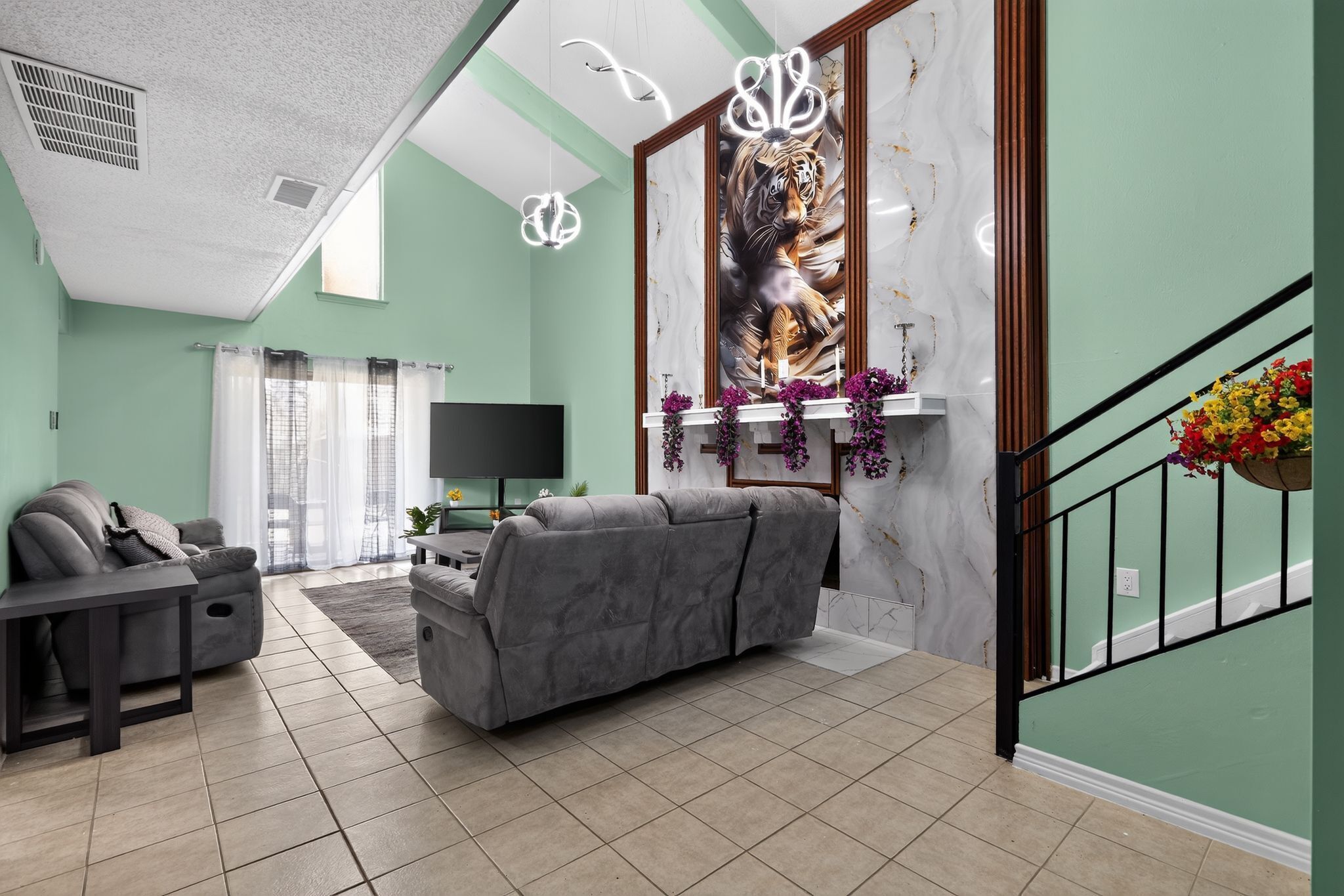 A living room with a gray sofa, a flat-screen TV, a fireplace with purple flowers, and a staircase with a flower basket.