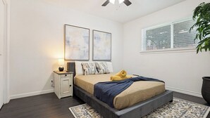 Ein Schlafzimmer mit Bett, Nachttisch, Lampe, Kunstwerk, Fenster und Deckenventilator.
