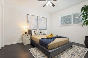 Ein Schlafzimmer mit Bett, Nachttisch, Lampe, Kunstwerk, Fenster und Deckenventilator.