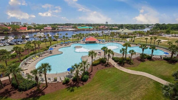 A large, winding pool surrounded by palm trees and lounge chairs, with a marina and buildings in the background. - Easy Breezy at Barefoot Resort w Amazing Pool (North Myrtle Beach)