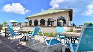 An outdoor patio with blue chairs and tables, a building with arched windows, and a clear blue sky. - Easy Breezy at Barefoot Resort w Amazing Pool (North Myrtle Beach)