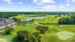 A golf course with a clubhouse, a lake, and surrounding forest. - Easy Breezy at Barefoot Resort w Amazing Pool (North Myrtle Beach)