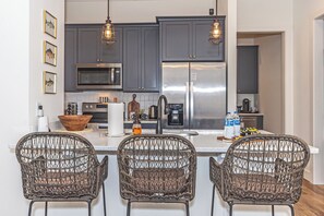 A modern kitchen with dark cabinets, stainless steel appliances, wicker chairs, and pendant lighting.