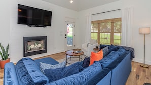 Ein Wohnzimmer mit einem großen, blauen Sofa, einem an der weißen Wand montierten Flachbildfernseher, einem Kamin und einem großen Fenster mit Jalousien.