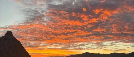 Una construcción con techo de paja, un área cercada y un paisaje montañoso bajo un cielo al atardecer.
