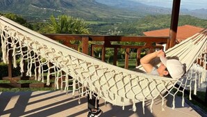 A person lying in a hammock on a terrace with a view of mountains and greenery.