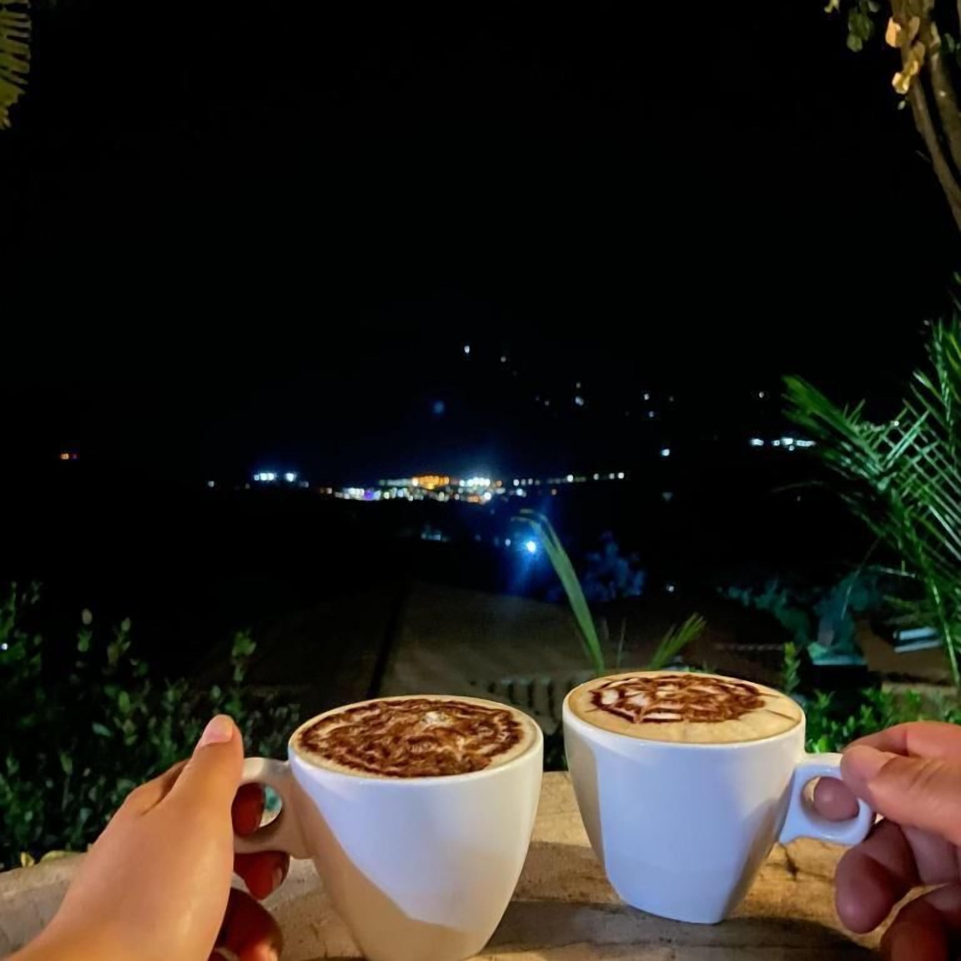Dos tazas blancas con arte latte en un balcón con vista a la ciudad iluminada de noche.