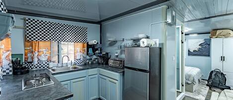 Eine kompakte Küche mit Spüle, Herd und Kühlschrank, ein kleiner Essbereich mit Tisch und Stühlen sowie ein Schlafzimmer mit Bett und Koffer.