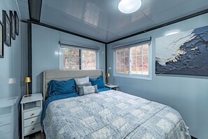 Een slaapkamer met een bed, twee ramen, een nachtkastje en een wandlamp.