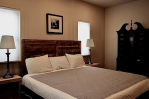 Un dormitorio con cabecera de madera, una cama con ropa de cama blanca, dos mesitas de noche con lámparas, un cuadro en la pared y un armario de madera oscura.