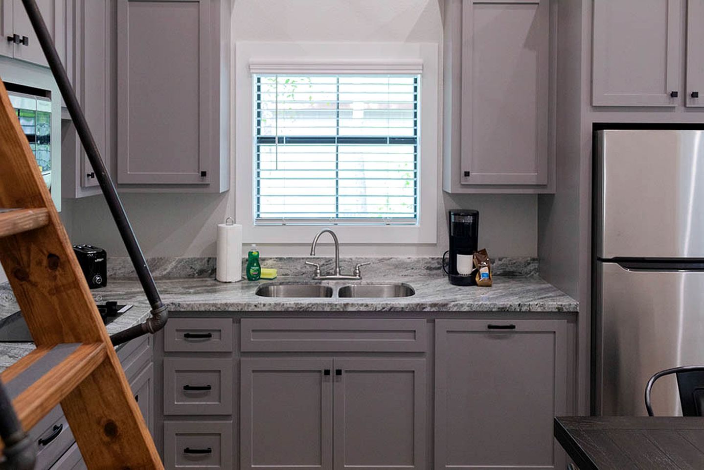 A kitchen with a stainless steel refrigerator, a coffee maker, and a window with blinds.