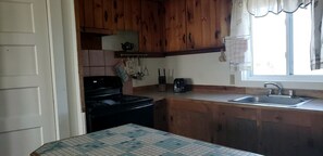 A kitchen with wooden cabinets, a stove, a sink, and a window with curtains. - Coastal Cottage Retreat with Direct Beach Access in North Truro, Massachusetts (North Truro)