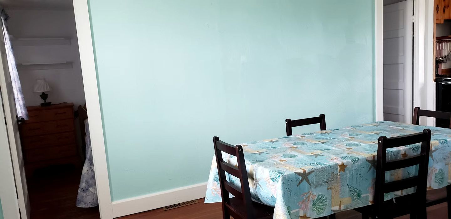 A dining area with a table covered by a floral tablecloth, two wooden chairs, a wooden cabinet, and a lamp on a nightstand.