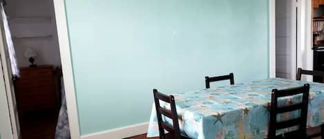 Un comedor con una mesa cubierta por un mantel floral, dos sillas de madera, un armario de madera y una lámpara en una mesita de noche.