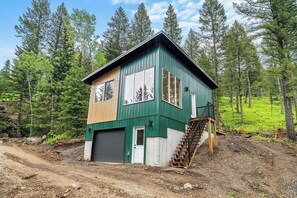 A two-story cabin with a front garage, a wooden staircase, and a dirt driveway.