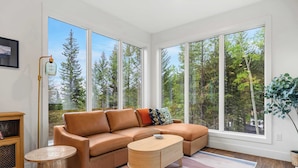 A modern living room with a brown leather sofa, a round coffee table, and large windows offering a view of trees.