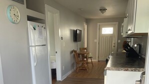 Una cocina con refrigerador, una mesa de comedor con sillas, un televisor montado en la pared y un cubo de basura.