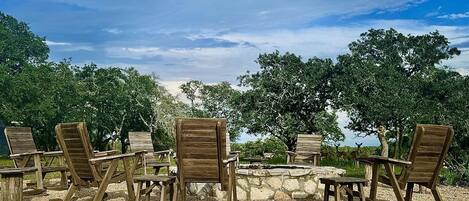 Outdoor seating with a stone fire pit, surrounded by trees and a clear sky.