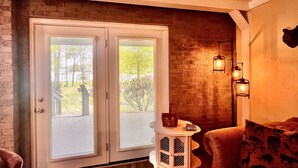 A room with a brick wall, a white door with glass panels, a small white table with a lamp, and a sofa with patterned cushions.