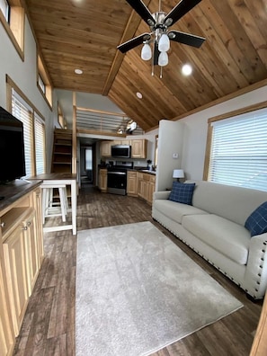 Un espacio habitable compacto con kitchenette, un sofá, ventilador de techo y una escalera que lleva a un nivel superior.
