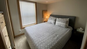 A bedroom with a bed, a nightstand, a lamp, a window with blinds, and a door.