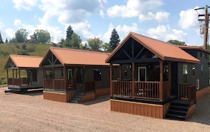 A row of cabins with wooden decks and metal roofs, set on a gravel ground with trees in the background.