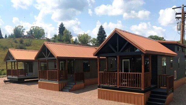 A row of cabins with wooden decks and metal roofs, set on a gravel ground with trees in the background.