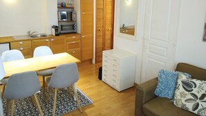 Cozinha compacta com micro-ondas, pia e armários de madeira. Há uma mesa de jantar com cadeiras e um pequeno armário branco. Uma sofá marrom com almofada estampada está na sala de estar.