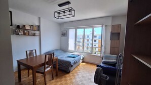 Una habitació petita i ben il·luminada amb un llit, una taula, una cadira i una finestra que dona vistes a edificis.