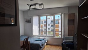 Habitació petita i lluminosa amb un llit, un escriptori i una cadira. Hi ha una finestra amb vista a edificis.