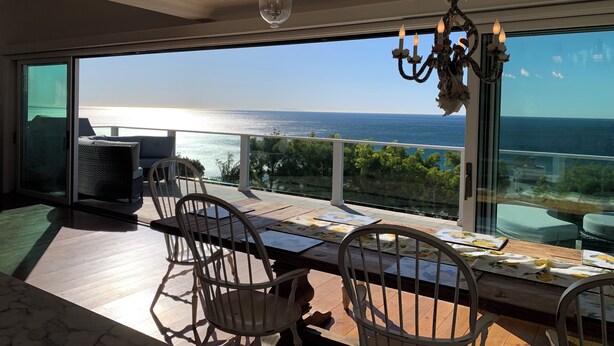 Uma varanda com vista para o mar, uma mesa com cadeiras e um lustre.