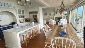 Uma cozinha espaçosa com uma ilha central, banquetas brancas e um espaço de jantar com mesa de madeira e cadeiras brancas.