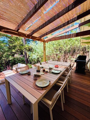 An outdoor dining area with a wooden table set for four, surrounded by chairs, under a pergola with a slatted roof. - Le Canon – Beautiful, renovated villa in the Landes region with a pool – Sleeps 10 – Beach within walking distance (Lège-Cap-Ferret)