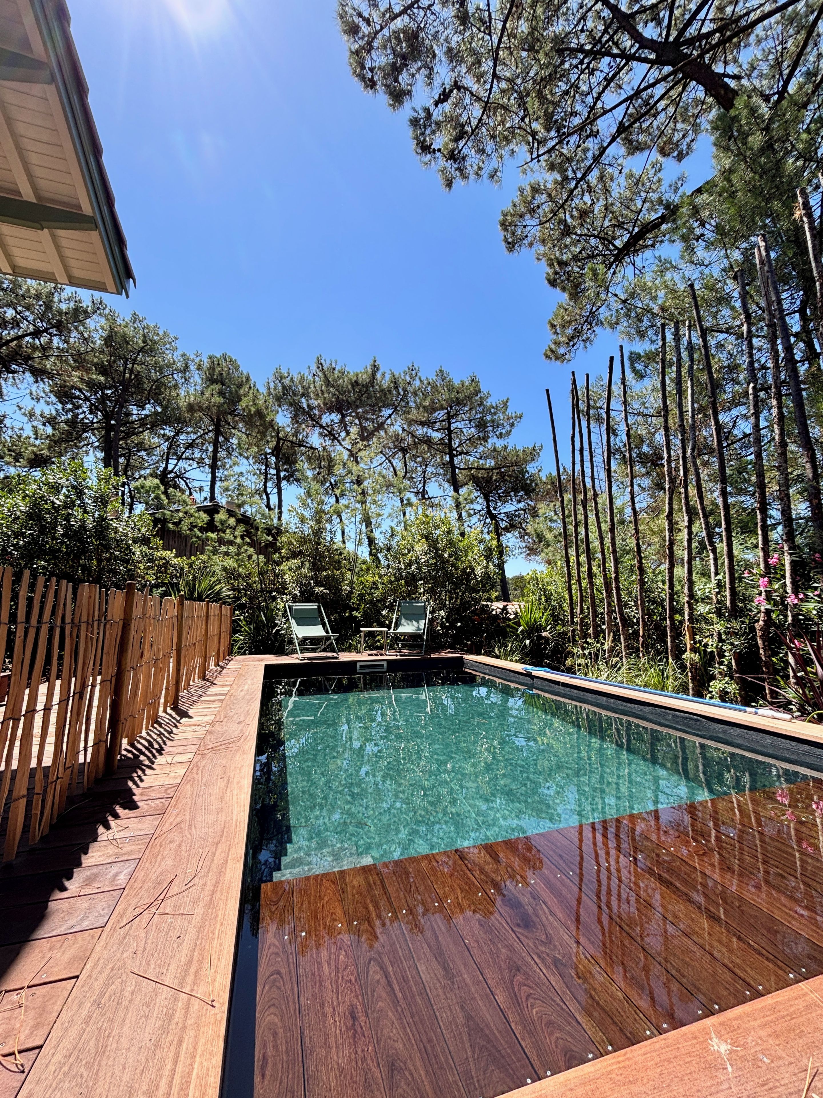 A swimming pool with a wooden deck, surrounded by trees and a wooden fence.